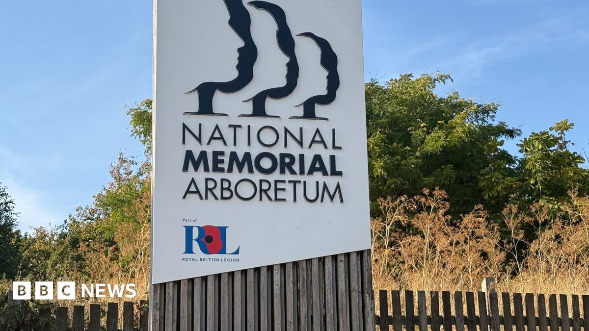 A large white sign that has the words "National Memorial Arboretum" on it with in smaller lettering "Part of Royal British Legion'. In the background is greenery and a fence.