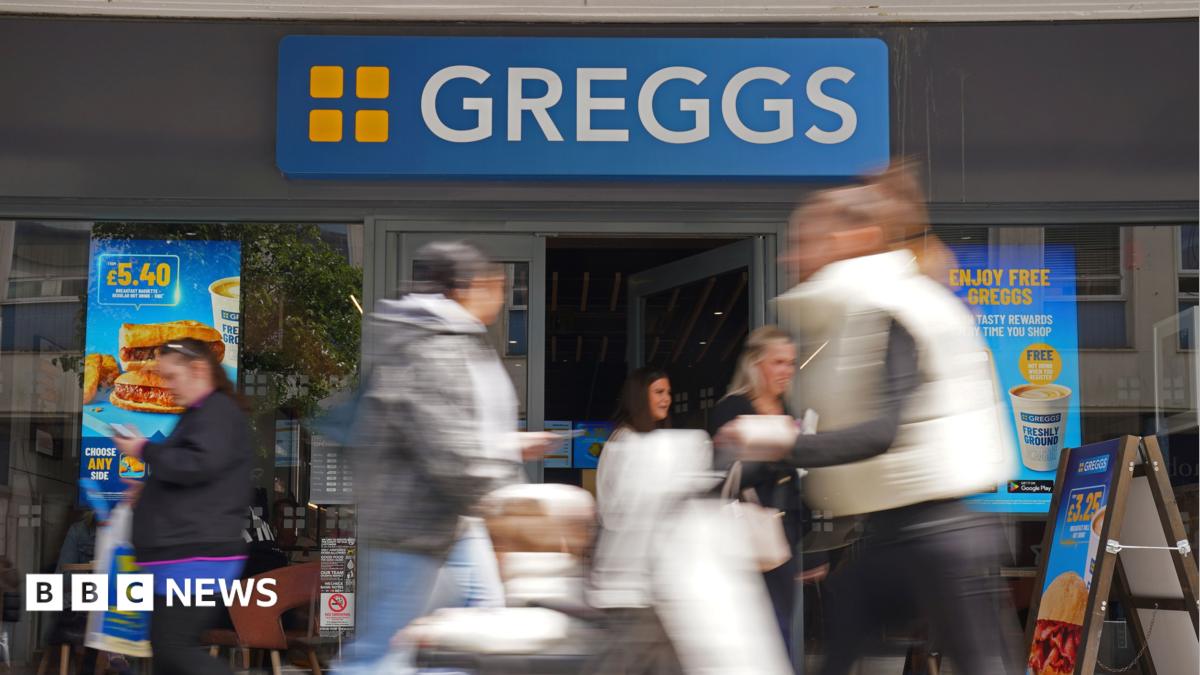 A stock image of a Greggs shop. Blurred figures of shoppers are portrayed walking past the shop and coming out of the store's door. There are adverts for their products on the windows.