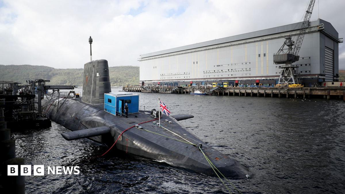 A submarine with a union flag flying from it, with a large white maintenance building in the background