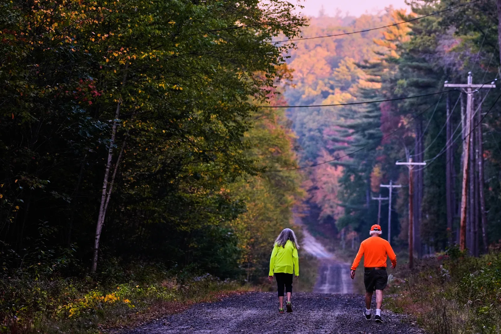A couple, wearing protective high-visibility clothing due to hunting season, take a walk at dawn, as trees of in the distance turn to fall foliage colors, Tuesday, October 7, 2025, in Chester, New Hampshire.
