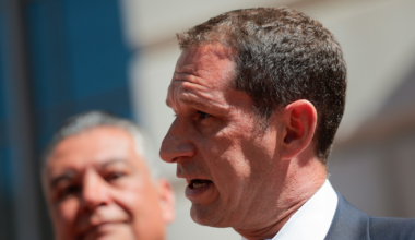 A man in a suit and blue tie speaks into multiple microphones, while another man in the background listens attentively.