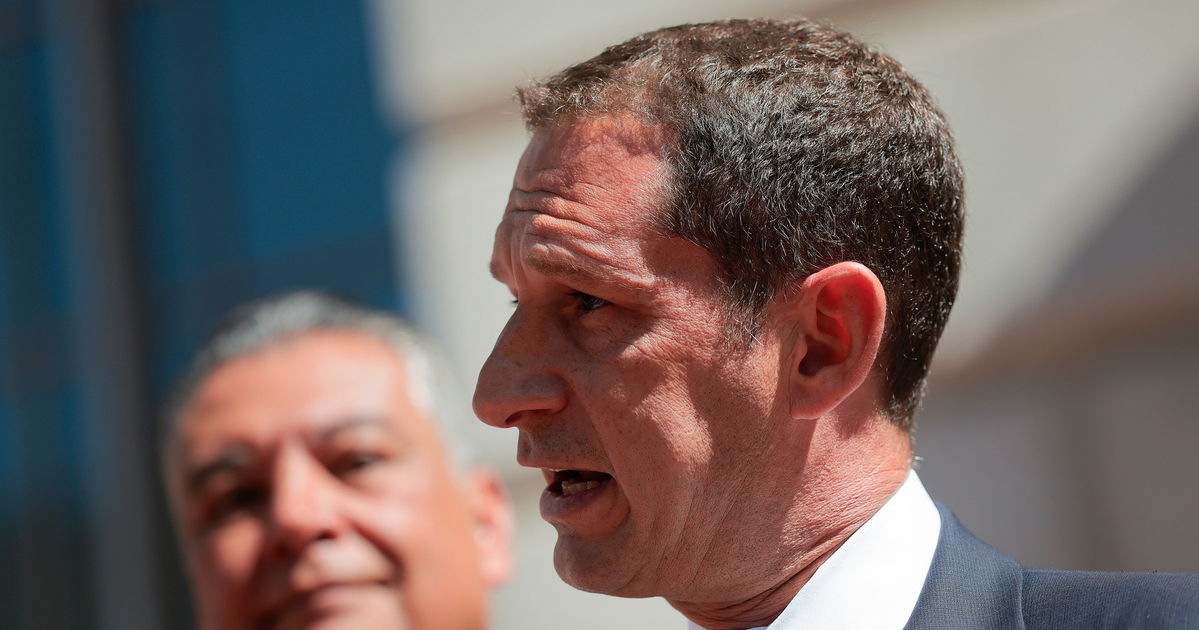 A man in a suit and blue tie speaks into multiple microphones, while another man in the background listens attentively.