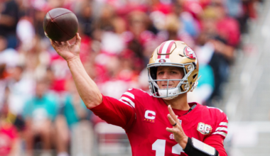 A football player in a red San Francisco 49ers jersey, number 13, throws a pass during a game with blurred spectators in the background.