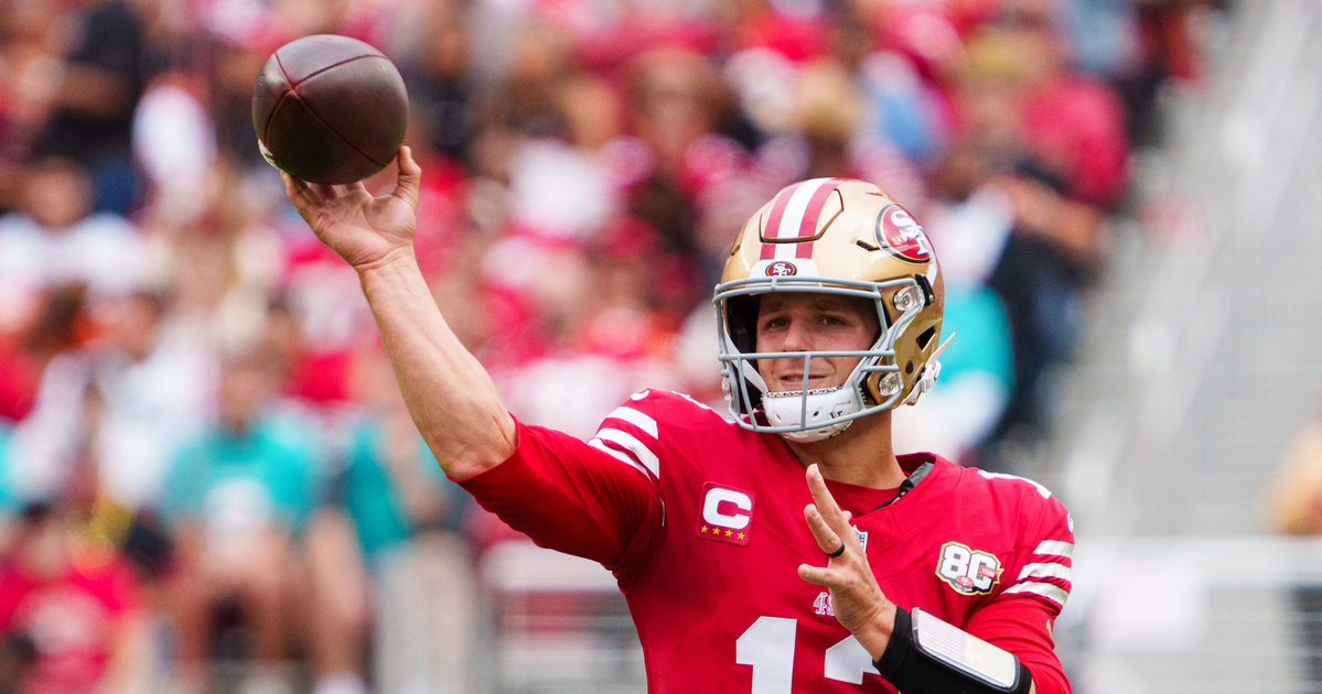 A football player in a red San Francisco 49ers jersey, number 13, throws a pass during a game with blurred spectators in the background.