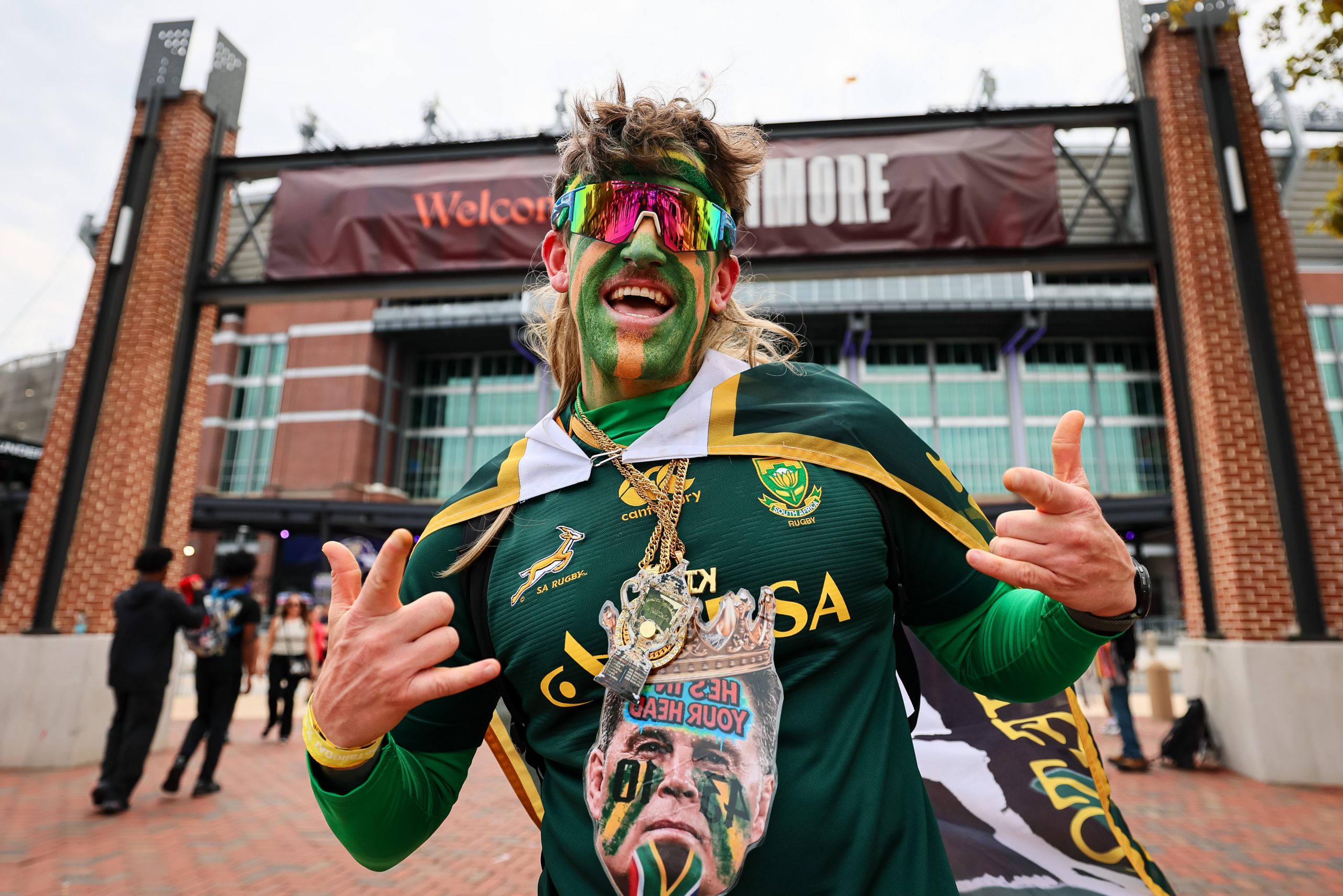 A smiling South Africa rugby supporter poses outside M&T Bank Stadium in Baltimore while wearing green and gold face paint, mulitcolored sunglasses and a South Africa rugby banner as a cape.