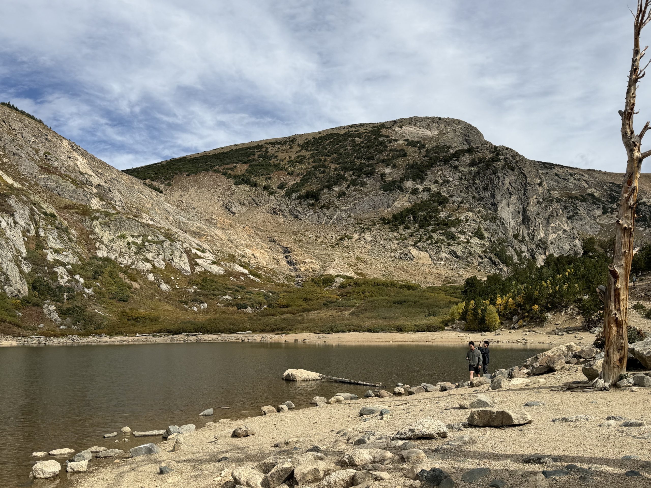 A rocky shoreline borders a small lake with people standing near the water, surrounded by mountains under a partly cloudy sky at St. Mary’s Glacier.