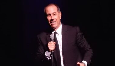 Jerry Seinfeld smiles while holding a microphone during a stand-up comedy performance.