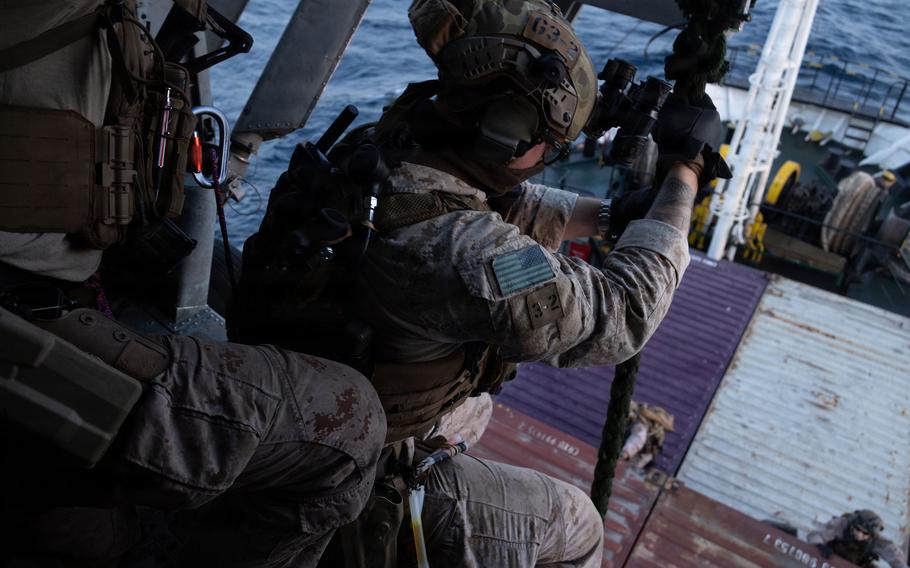 A close-up view of a Marine in camouflage combat gear and black bullet-proof vest, seen from behind, standing on a ramp at the back of a ship and preparing to board an adjacent ship.