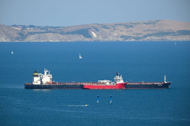 PORTLAND, ENGLAND - AUGUST 10: Two hydrofoil kite surfers pass the offshore tug and supply ship FS Aries which alongside the oil tanker Smyrtos as it continues to sit outside Portland harbour, on August 10, 2026 in Portland, United Kingdom. The oil tanker operated as part of Russia's shadow fleet and was intercepted by British Forces in the English Channel in June. (Photo by Finnbarr Webster/Getty Images)