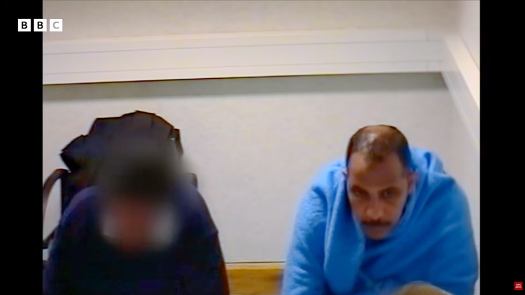 A man is seen in a plain white room while another figure with a blurred face sits to his right.