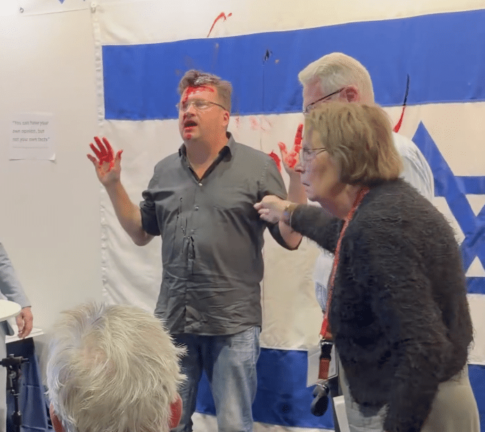 Man with red paint on his face and hands stands in front of an Israeli flag at the Gothenburg Book Fair as people gather around him.
