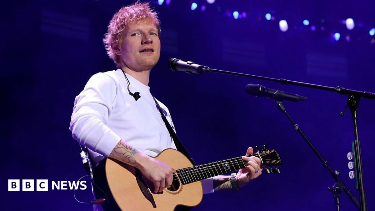 Ed Sheeran, in a long sleeved white T-shirt, holds a guitar and sings into a microphone. He is looking slightly off to one side.