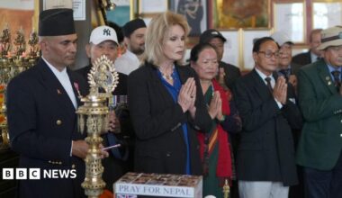 A woman with long, light hair stands with her hands together as though in prayer. She stands in a row of men and women of Nepalese appearance, many with their hands together.