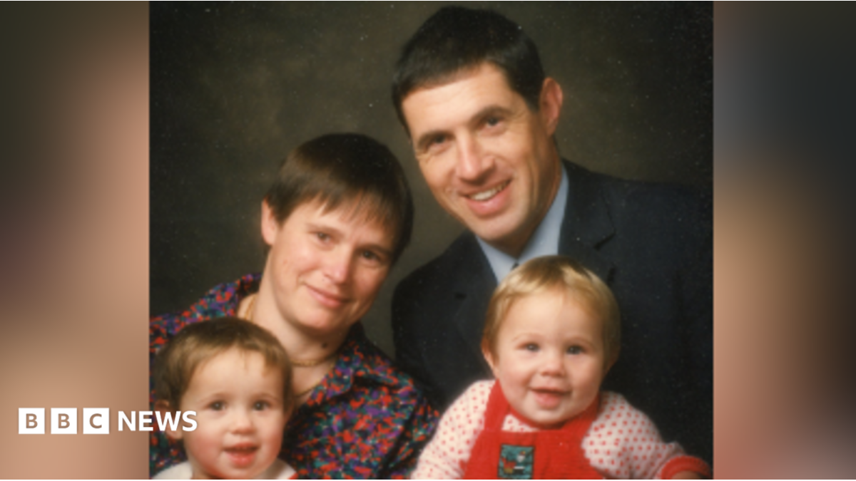 A family photograph of Helen and Clive with their two young children. Helen has short brown hair and wears a red and blue patterned top. Clive wears a suit with a pale blue shirt. Helen holds a toddler who is wearing a red jumper while Clive holds a baby wearing red dungarees.