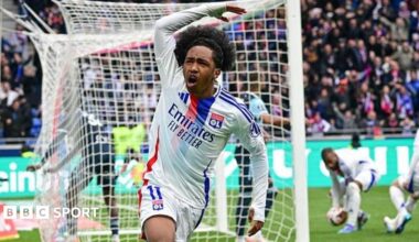Malick Fofana, wearing Lyon's white kit with a red-and-blue stripe, celebrates scoring a goal by waving his right arm in the air