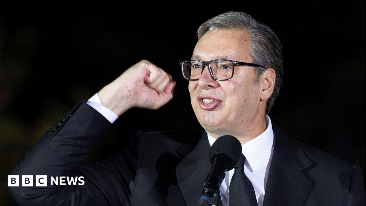 A head shot of Aleksandar Vucic looking off-camera giving a speech while waving his fist in the air. He is wearing a black suit and tie with white shirt.
