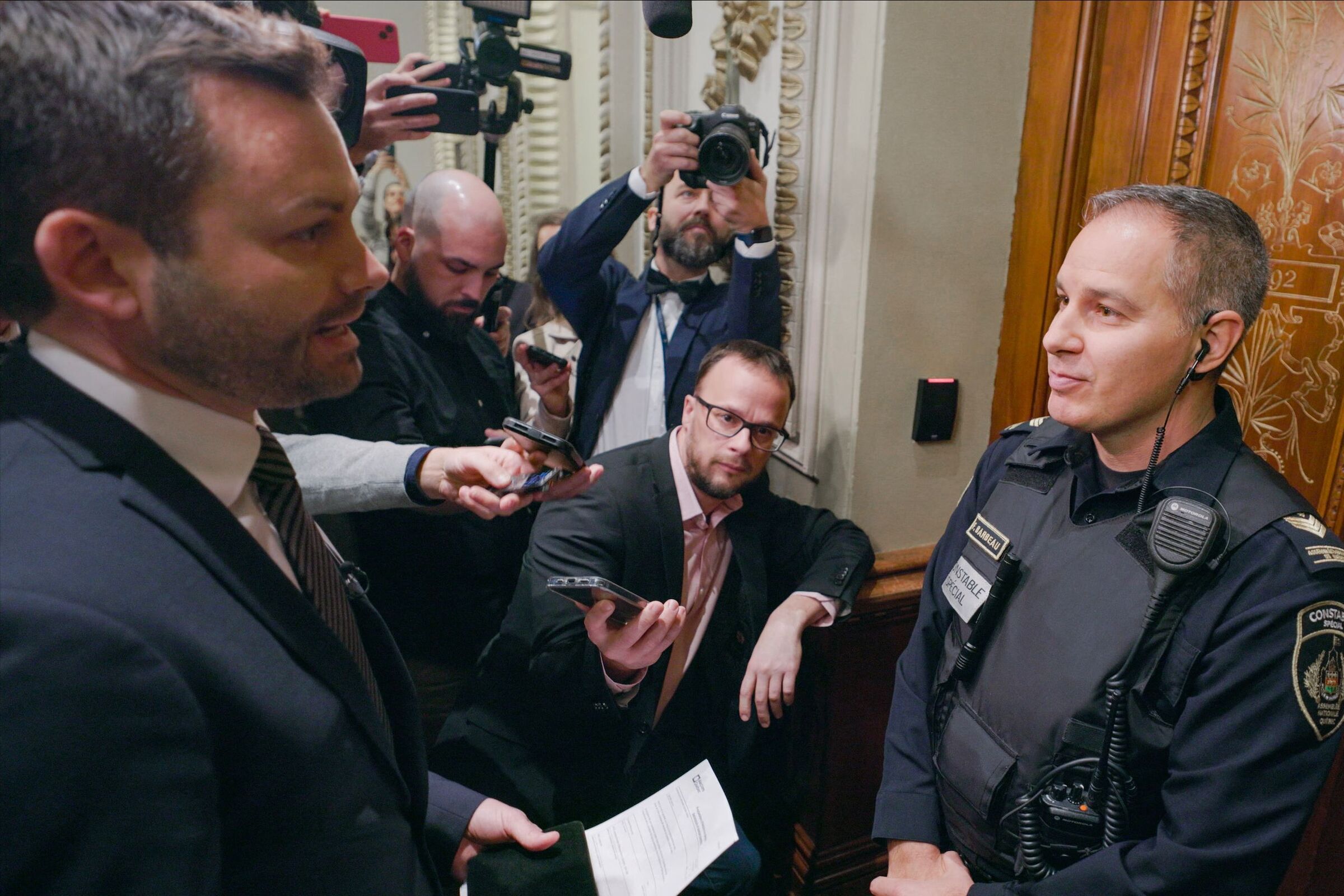 A police officer stands between Paul St-Pierre Plamondon and a door as journalists watch from the side.
