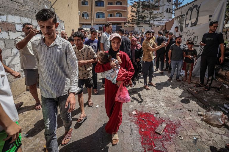 A Palestinian woman carries a girl past a blood stain on the ground after an Israeli drone strike on a cafe in al-Bureij camp, central Gaza Strip, on September 16, 2026