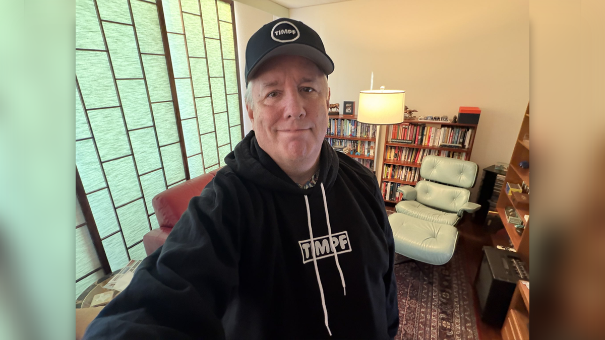 Andrew Wimsatt in a selfie photo taken inside a home library while wearing a black hoodie and ballcap