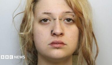 A mugshot of a young woman with blonde hair. It tied back in a messy ponytail. She has her lip, eyebrow, nose and the bridge of her nose pierced with metal bars and has a neutral expression.