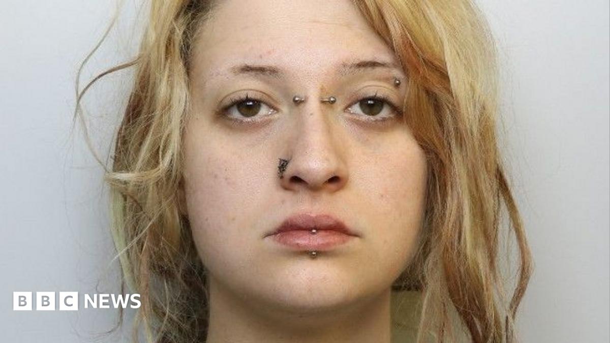 A mugshot of a young woman with blonde hair. It tied back in a messy ponytail. She has her lip, eyebrow, nose and the bridge of her nose pierced with metal bars and has a neutral expression.