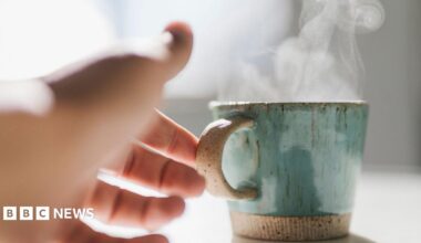 A woman reaches her hand out to pick up a steaming hot mug which is shiny and green-coloured