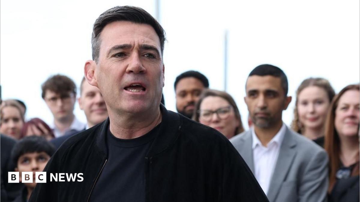 Andy Burnham is wearing a dark jacket over a dark top. He is stood in front of a crowd of people and is speaking.