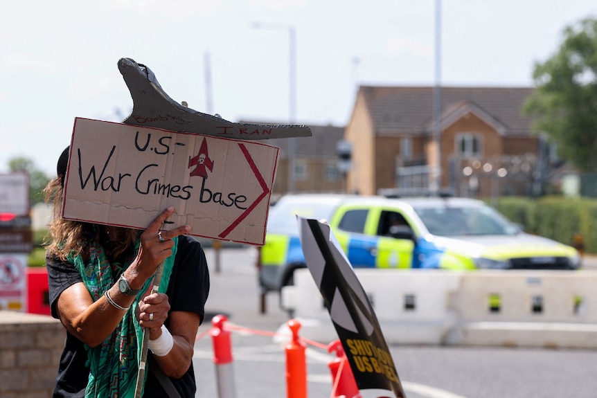 A person holds up a cardboard protest placard saying "US War Crimes Base".