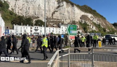 A group of people in black clothing wearing black balaclavas. A number of police officers are in the crowd.