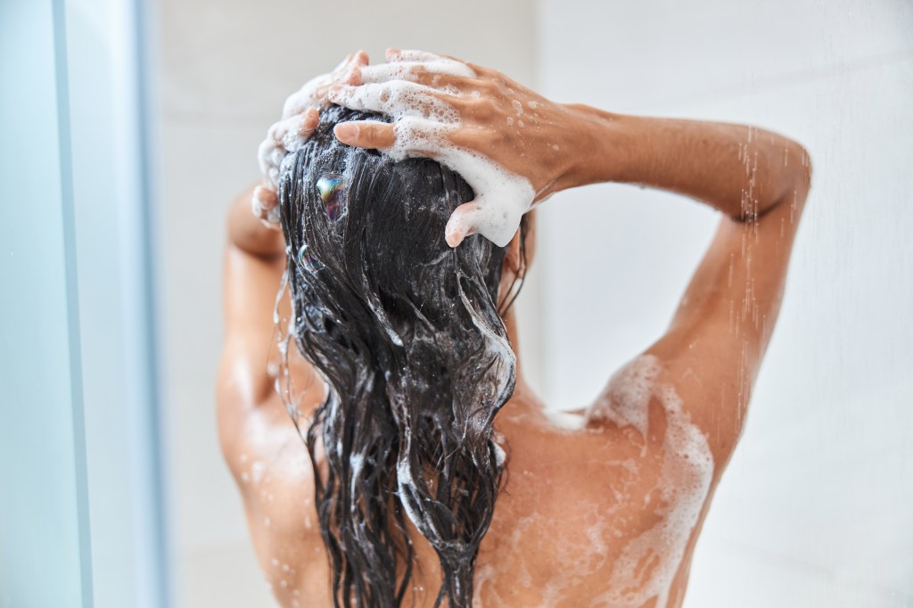 Back view of a woman washing her long, dark hair with shampoo in the shower.