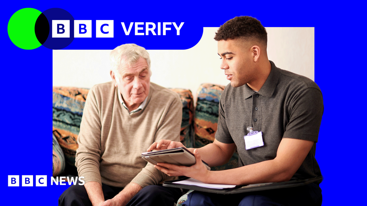 A senior man sat down speaking to a younger adult wearing a lanyard and holding a tablet.