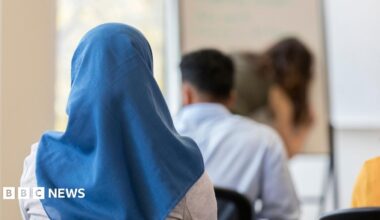 A stock image of a girl wearing a blue headscarf. She is sitting down in a classroom, facing with her back towards the camera.
