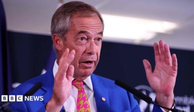 Nigel Farage speaking in front of two microphones, wearing a blue suit and a pink and yellow tie. He is wearing a union flag-patterned badge on his lapel.