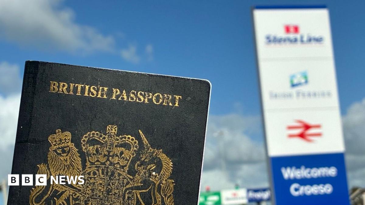 A dark blue British passport in the foreground with a sign welcoming people to Holyhead port, slightly out of focus in the background