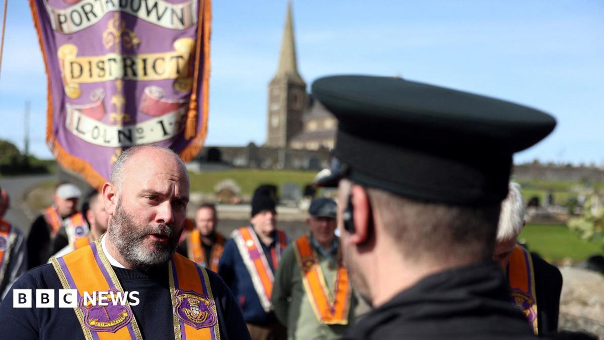 A man with a greying beard speaks to a PSNI officer. He wears an Loyal Orange Order collarette. In the background, other Orangemen stand and the church is also seen in the background.