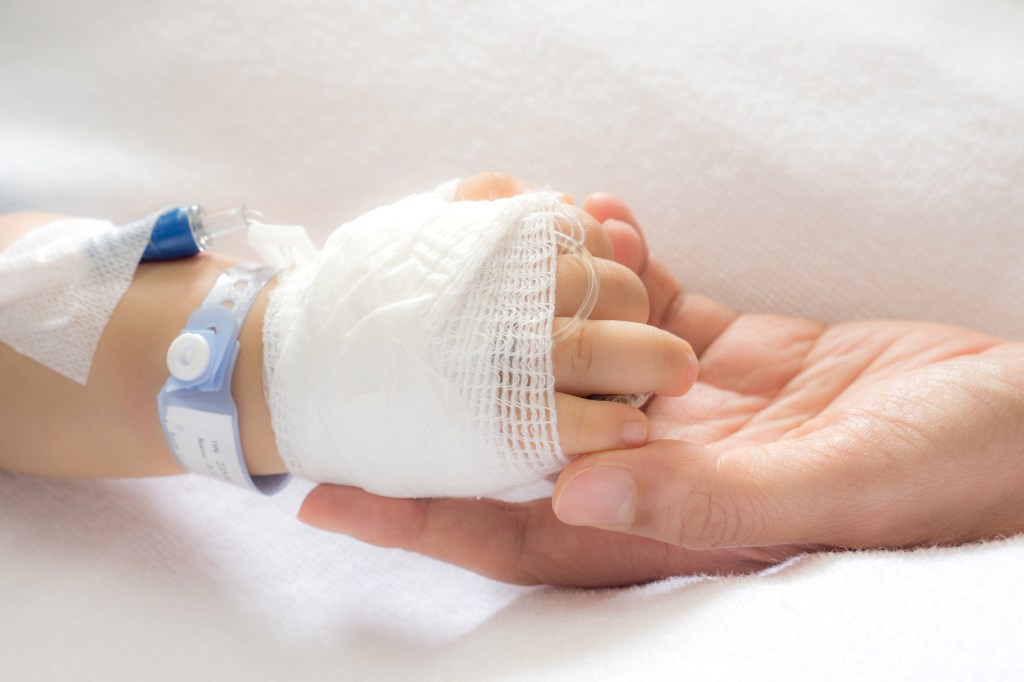 A child's bandaged hand with an IV drip and hospital wristband is gently held by an adult's hand.