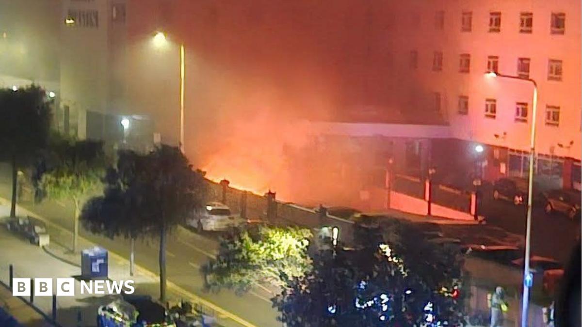 A fire in the basement of a hotel. Flames can be seen from below the building, and emergency services can be seen in the foreground.