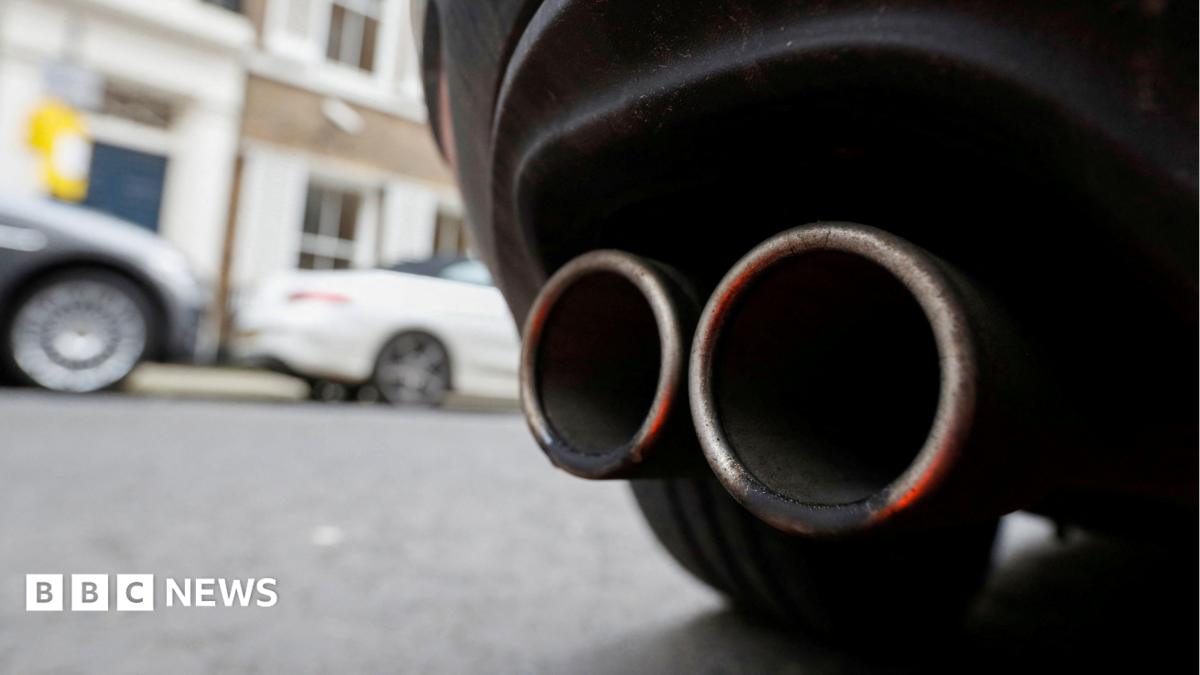 An exhaust pipe on the back of a car. A wheel can be seen, with two cars out of focus in the background