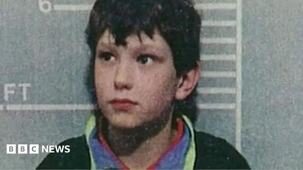 A police custody shot of a young Jon Venables, wearing a dark jumper, after his arrest. He is standing in front of a height chart showing he stands just over 4ft 6in tall.