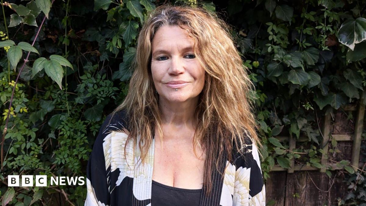 Helen with shoulder length wavy brown hair wearing a black vest and a black white and gold kimono style top standing in a garden with fence and foliage behind her