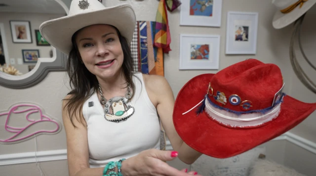Jen Lott, wearing a white cowboy hat, holds up a red embellished cowboy hat as she smiles at the camera