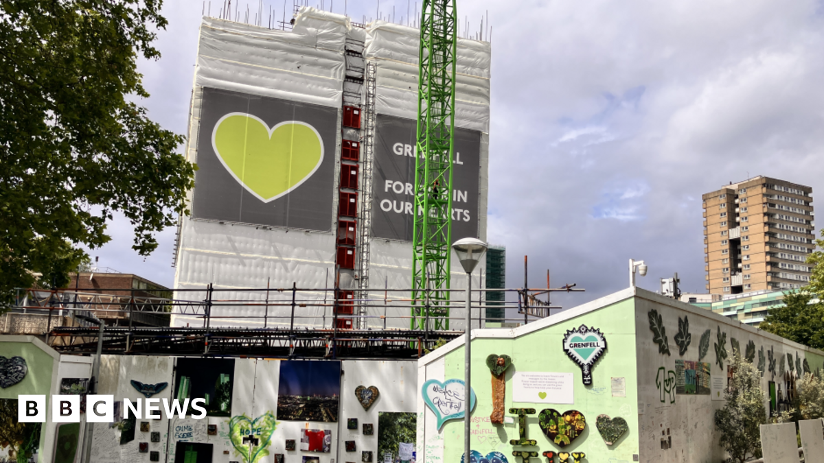 A partially demolished Grenfell Tower with a wall surrounding the area which is covered in tributes and drawings. The building is covered in a wrap which features a green heart.
