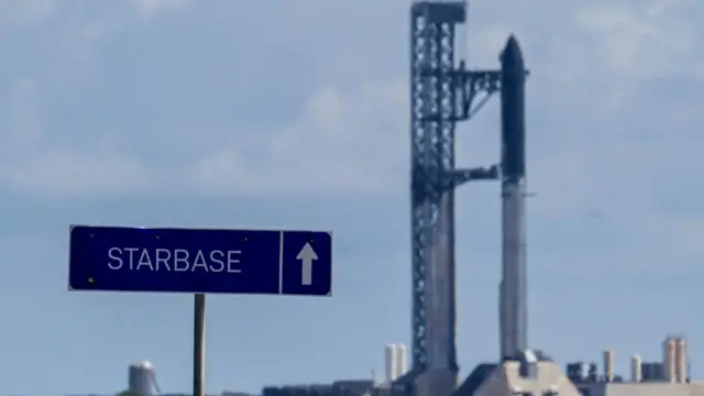 A rocket being supported by a huge metal structure in the distance, blurry. A blue sign in the foreground says Starbase with an arrow in the direction of the rocket and buildings that surround it
