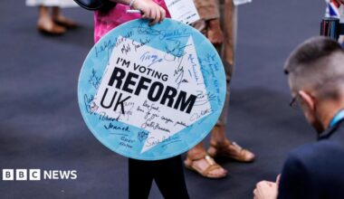 A woman holding a sign saying I'm Voting Reform UK enters the Reform conference. The sign has autographs on it from Reform politicians.