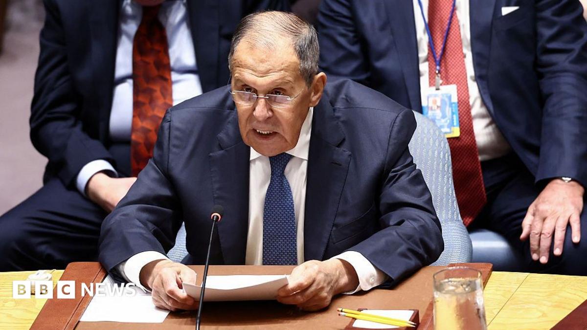 Sergey Lavrov wearing a navy suit, white shirt, and blue tie. He is sat behind a microphone with other diplomats in suits behind him. Lavrov is holding several documents in his hands, wears glasses, and has short grey hair.