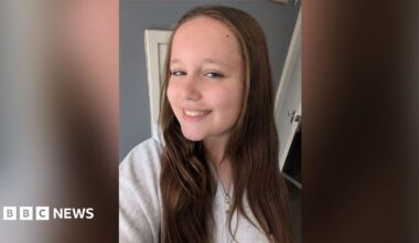 Lillia stands smiling to the camera whilst taking a selfie. She has brown hair which sits on her shoulders and is wearing a white top.
