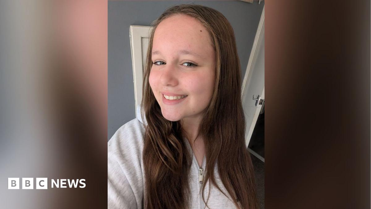 Lillia stands smiling to the camera whilst taking a selfie. She has brown hair which sits on her shoulders and is wearing a white top.