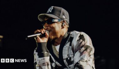 Jay-Z raps into a microphone while performing on stage. He wears a black and white cardigan and a New York Yankees hat.