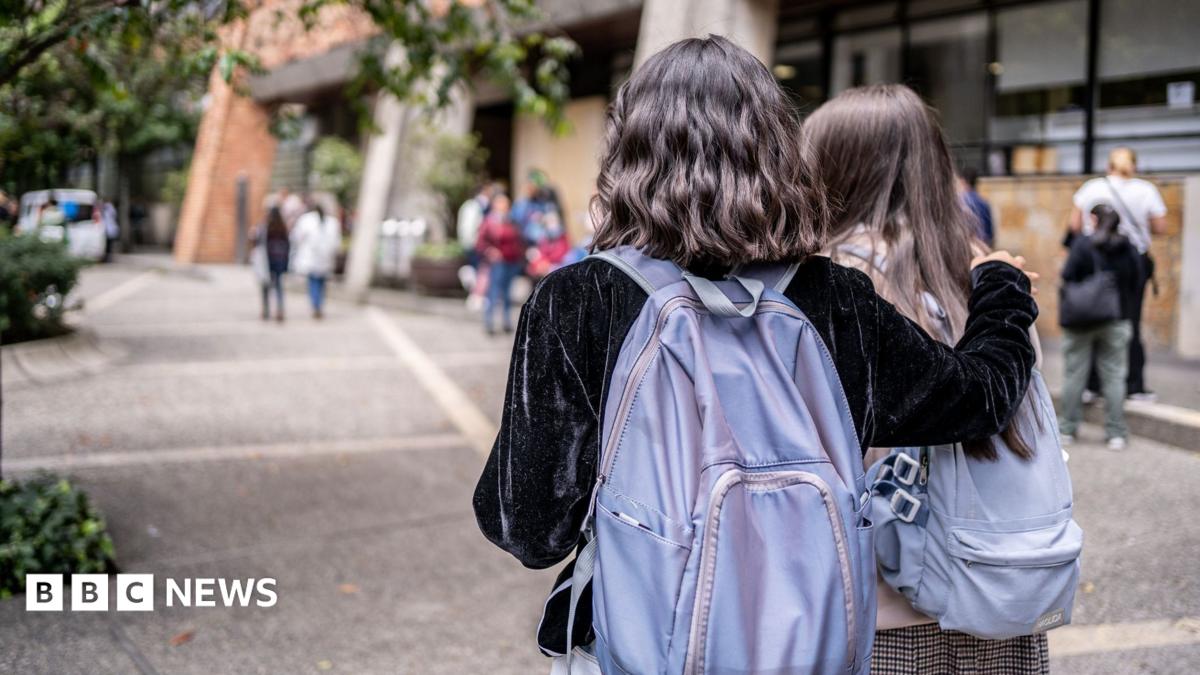 Two university students with purple backpacks walk across a university campus. Only the backs of their heads are visible. One has short wavy dark hair and wears a black velvet jacket. She has her right arm around the second student, who has long dark hair.