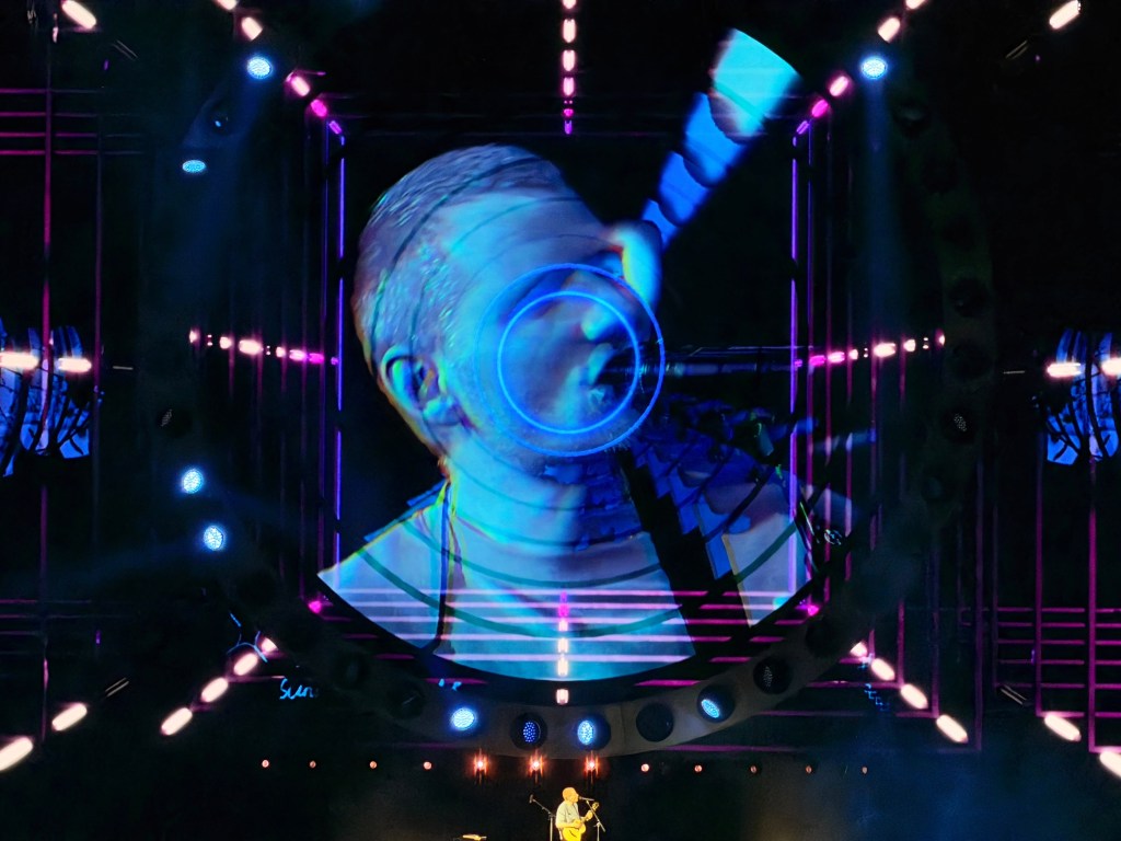 Ed Sheeran performing on stage with a large screen behind him displaying his face.